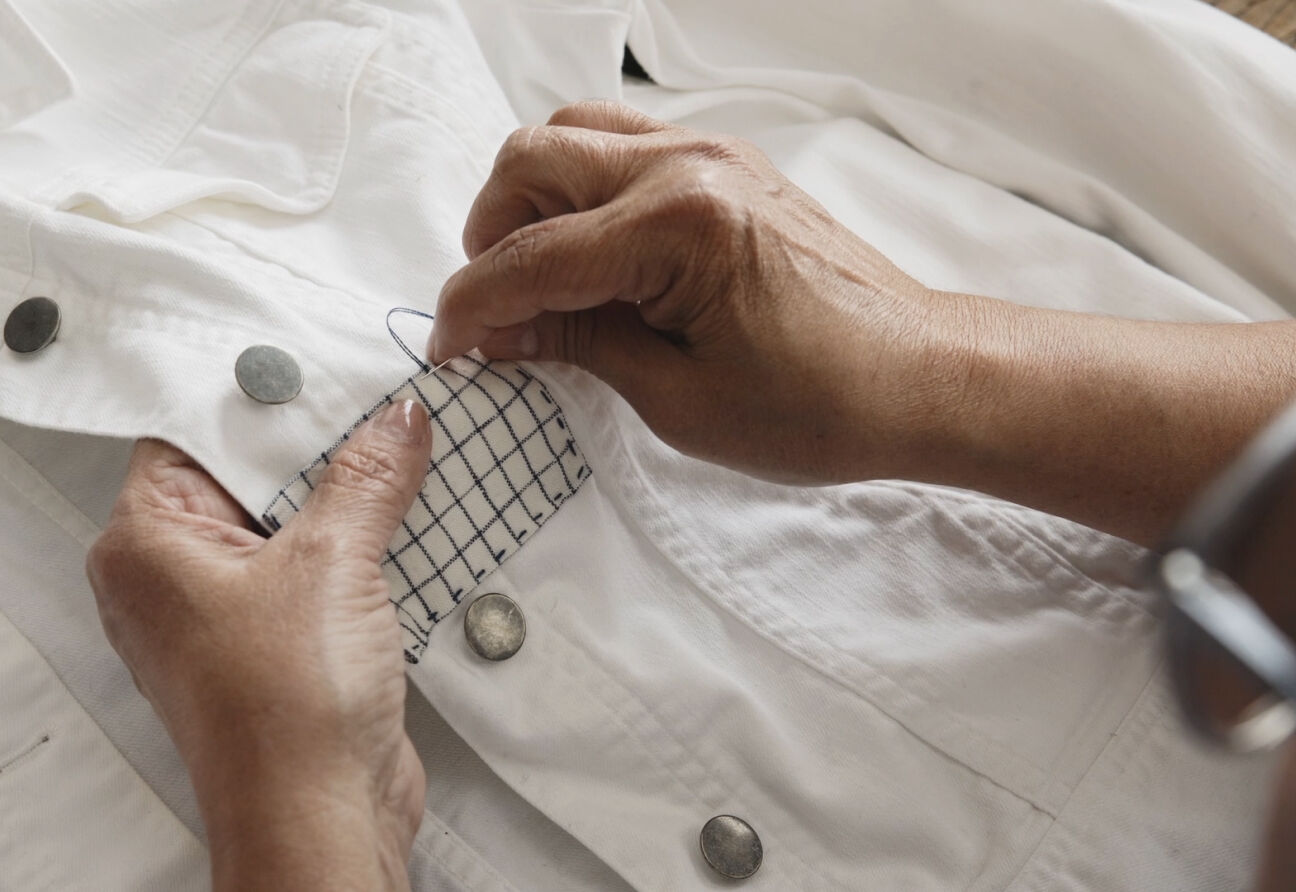 Picture of hands sewing a fabric patch on a torn white denim jacket.