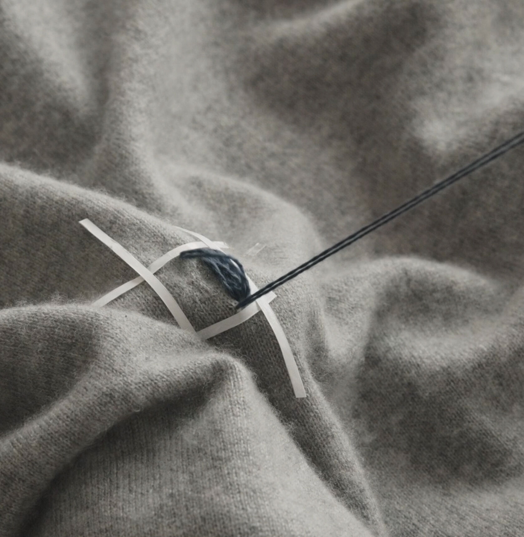 Close-up of hands stitching blue thread vertically on a mended gray sweater.