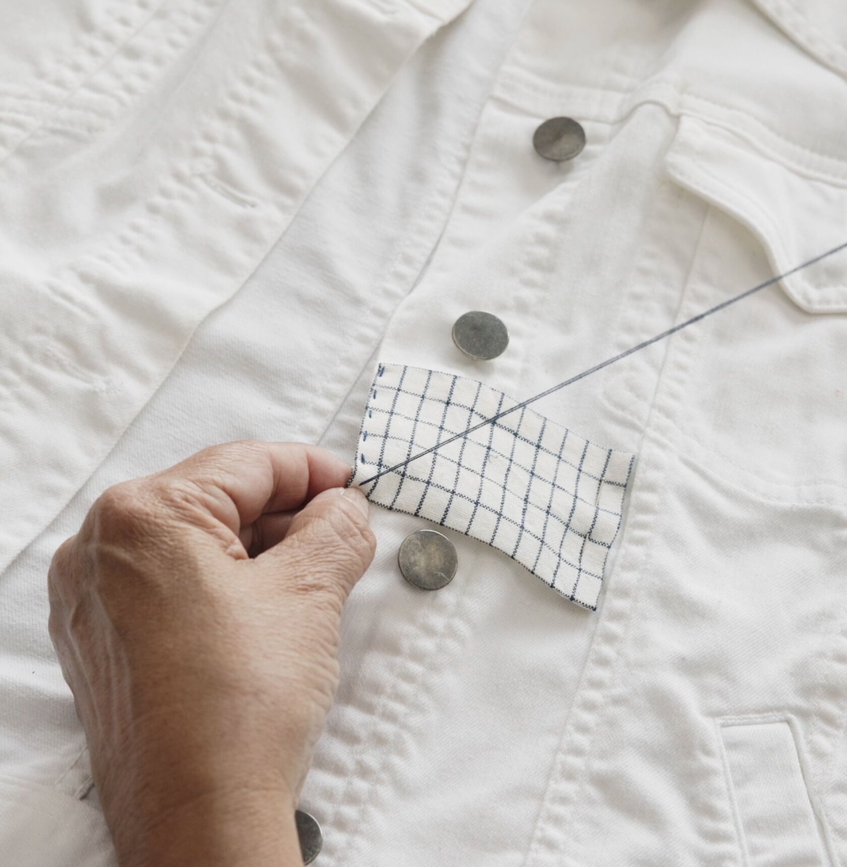 Hands stitching a patch on white denim jacket.