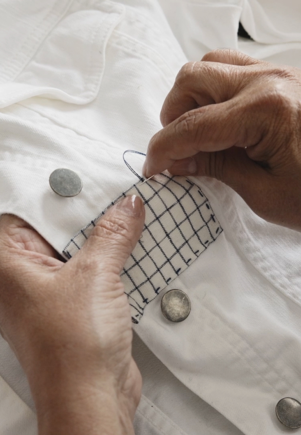 Picture of hands sewing a fabric patch on a torn white denim jacket.